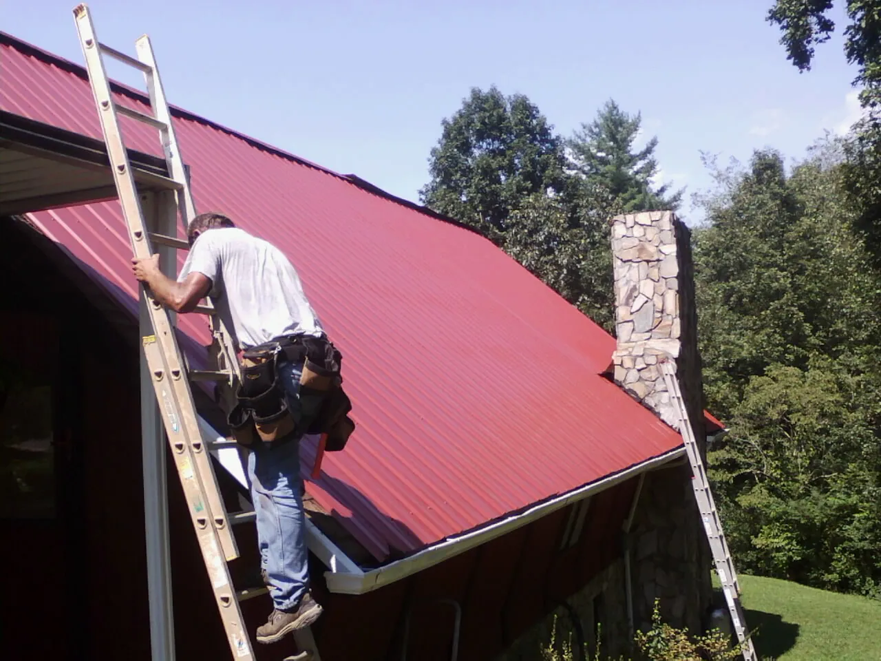 Roofer on a ladder beside a red metal roof and stone chimney — Soffit & Fascia Repair by Roofing Brothers in Salvisa